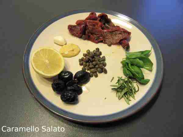 Spellare l'aglio, lavare il basilico e il rosmarino, prelevare gli aghi, sgocciolare i pomodori secchi e sciacquare dal sale i capperi, snocciolare le olive